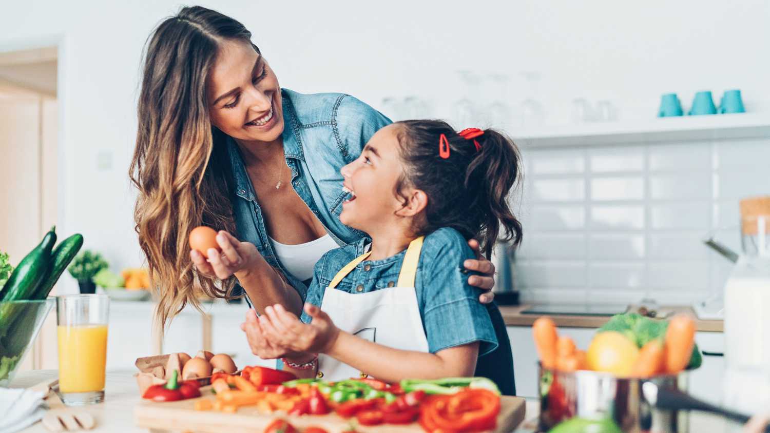 Uśmiechnięte mama z córką siedzą w kuchni przy stole pełnym warzyw i przygotowują zdrowy posiłek, bo zbilansowana dieta i zdrowy tryb życia to metody na to, jak zapobiegać paradontozie.