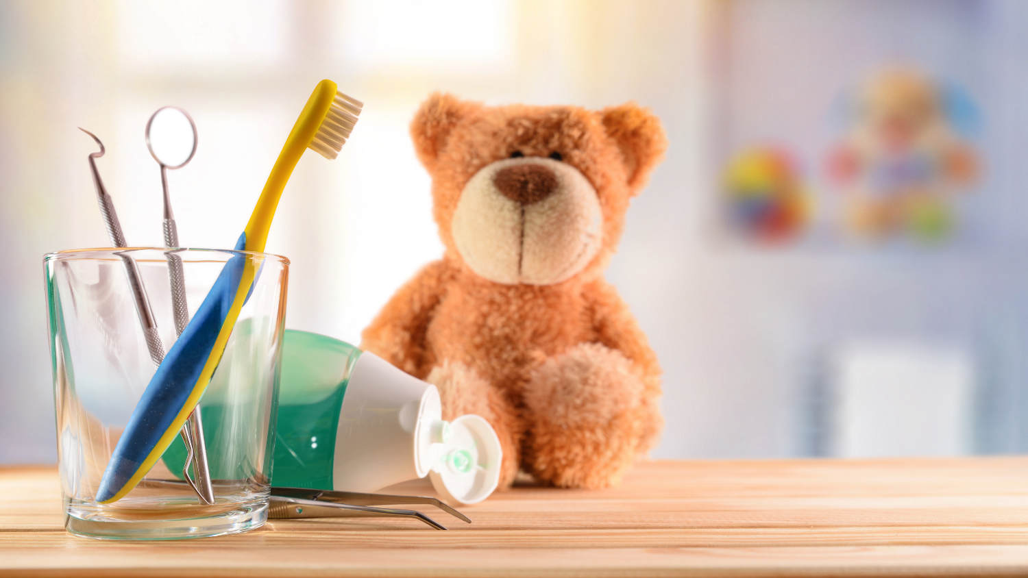 Pluszowy miś siedzi na blacie, a obok znajduje się kubek ze szczoteczką i lusterkiem dentystycznym oraz pasta do zębów, co symbolizuje leczenie zębów mlecznych mleczaków u dzieci.