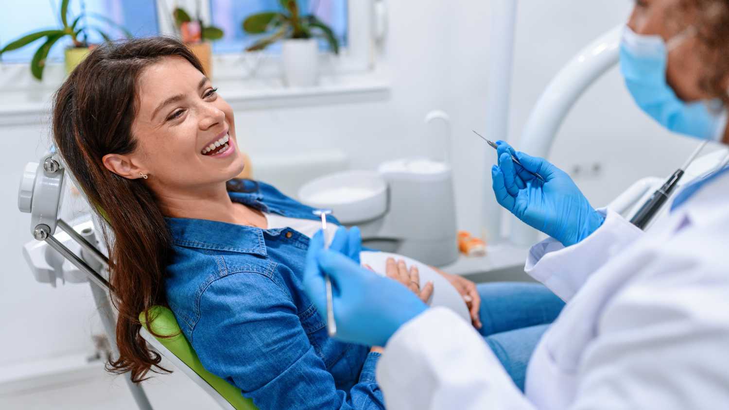 Kobieta w ciąży uśmiecha się i siedzi na fotelu dentystycznym, a dentysta wyjaśnia, jak wygląda kwestia ciąża a leczenie stomatologiczne.